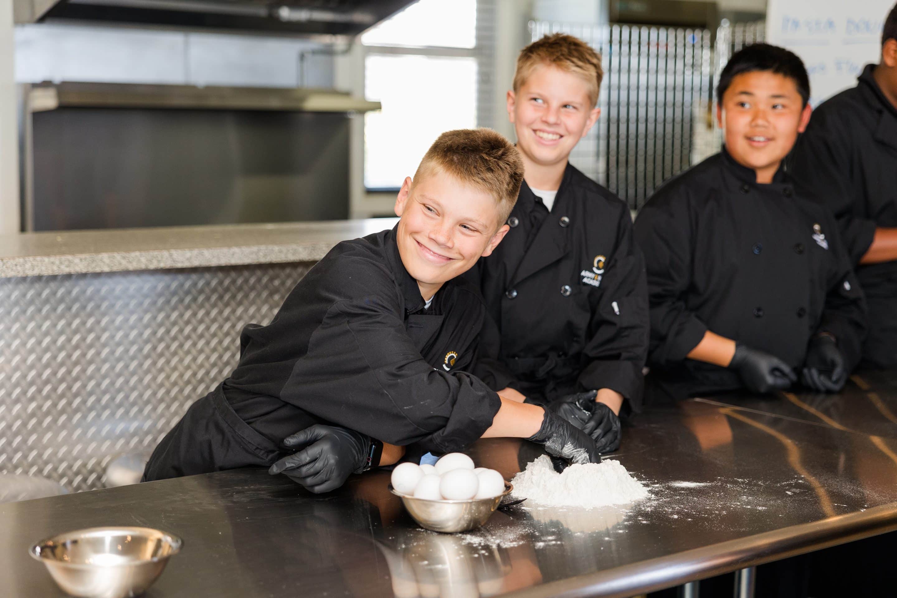 Four young people in black chef uniforms work together in a kitchen.